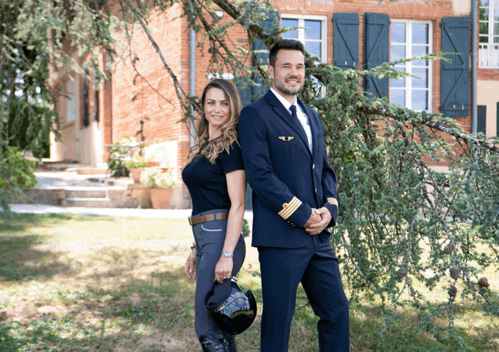 Portrait extérieur d'un couple : un pilote souriant en uniforme et une femme en tenue d'équitation tenant un casque noir, devant une bâtisse en briques.