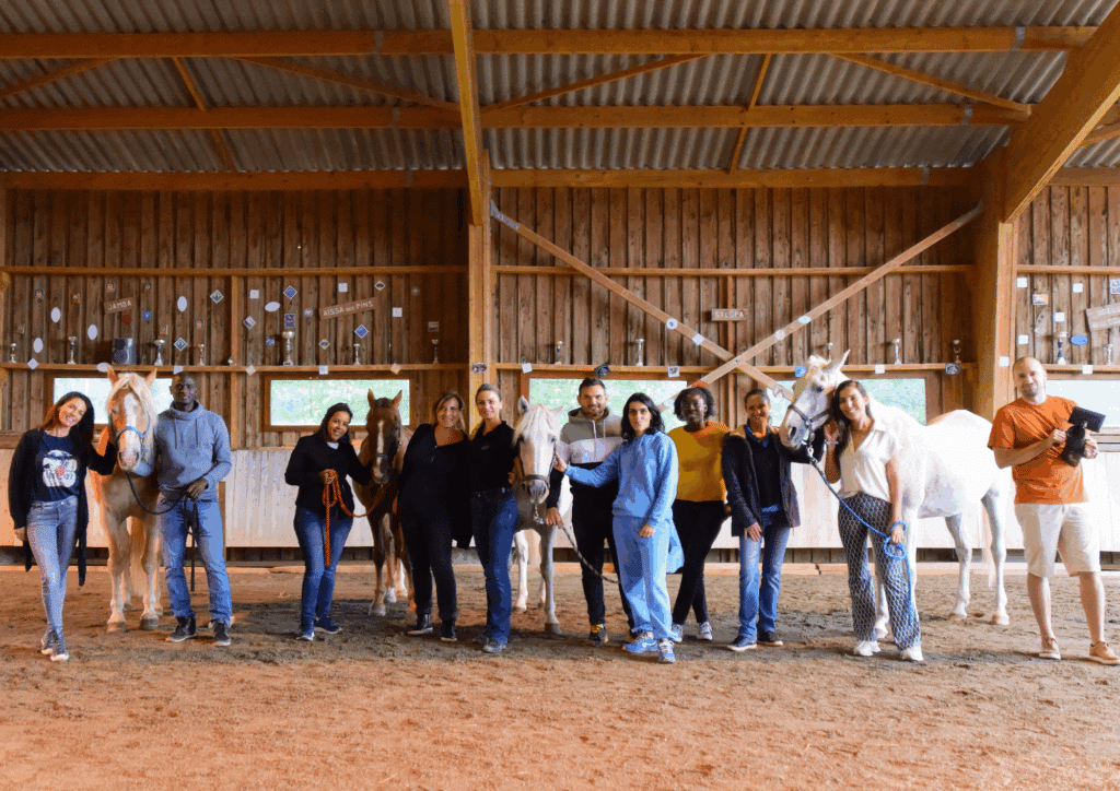 Groupe de 11 personnes souriantes avec quatre chevaux dans un manège intérieur en bois, affichant des trophées et des noms comme 'JAMBA' et 'AÏSSA DES PINS'.